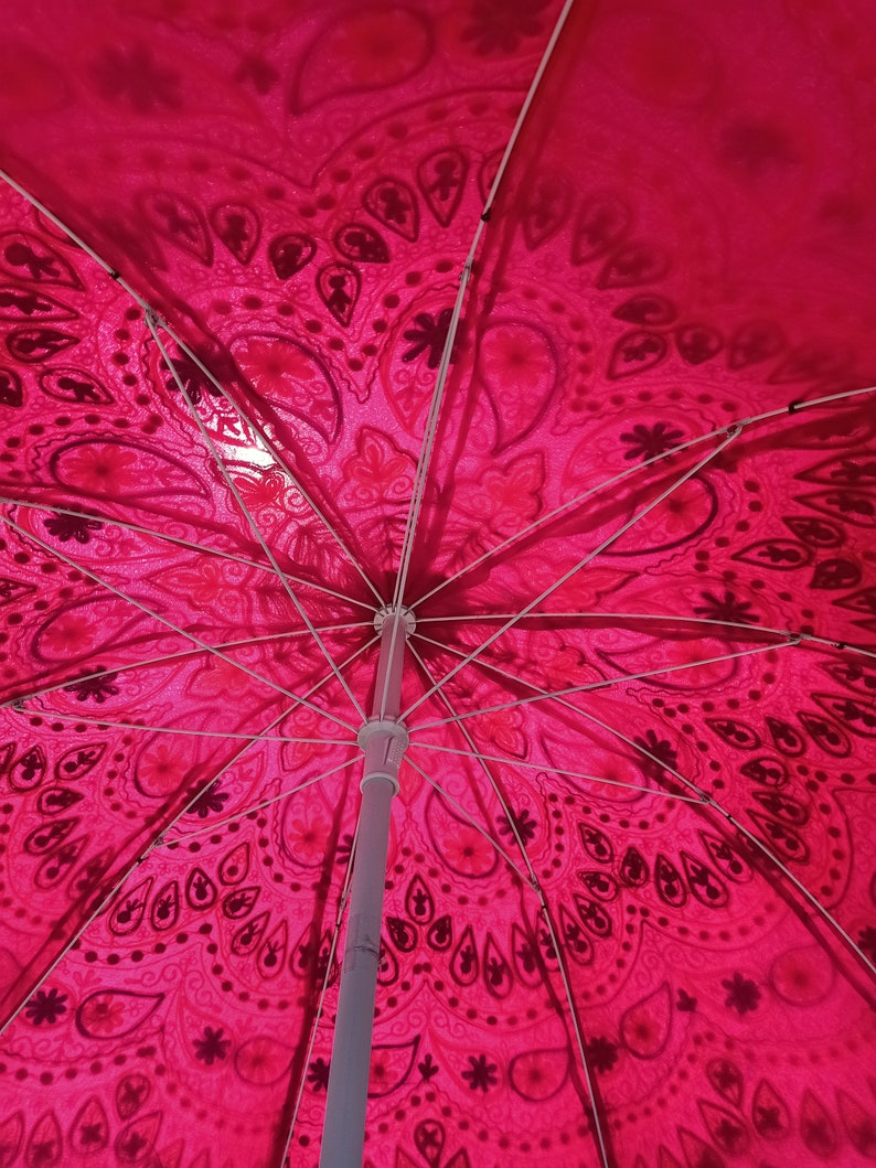 Może przedstawiać: R&oacute;żowy i biały parasol plażowy z wzorem i białą metalową ramą. Parasol jest otwarty, a wnętrze baldachimu jest widoczne.