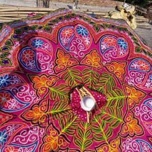 Może przedstawiać: R&oacute;żowy parasol z kolorowymi haftami przedstawiającymi wzory kwiatowe i paisley. Parasol jest wykonany z tkaniny i ma białe centrum.