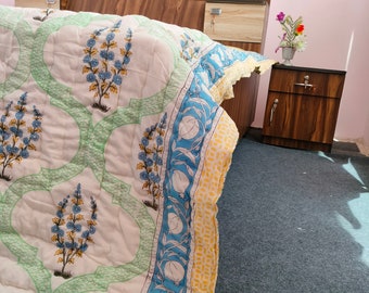 May include: A white and green quilt with a blue and yellow border. The quilt has a floral pattern and is folded on a bed with a wooden headboard. The headboard has a carved apple design.