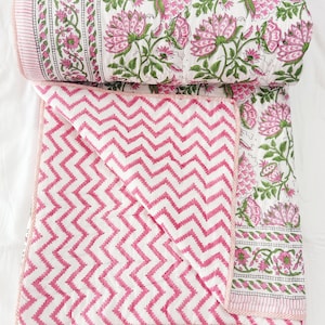 May include: A folded pink and white quilt with a chevron pattern and a rolled-up quilt with a floral design. The floral design features pink flowers and green leaves on a white background. The quilt has a pink border.