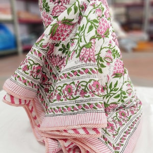 May include: A folded white quilt with a floral pattern in pink and green. The quilt features a border with a pink and white striped design. The floral pattern includes large pink flowers and green leaves.