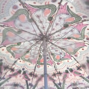 Może przedstawiać: Biały parasol plażowy z kolorowym, skomplikowanym wzorem. Wz&oacute;r zawiera kształty paisley, ptaki i elementy kwiatowe w odcieniach r&oacute;żu, zieleni i pomarańczy. Parasol ma biały słup i metalowe żebra.