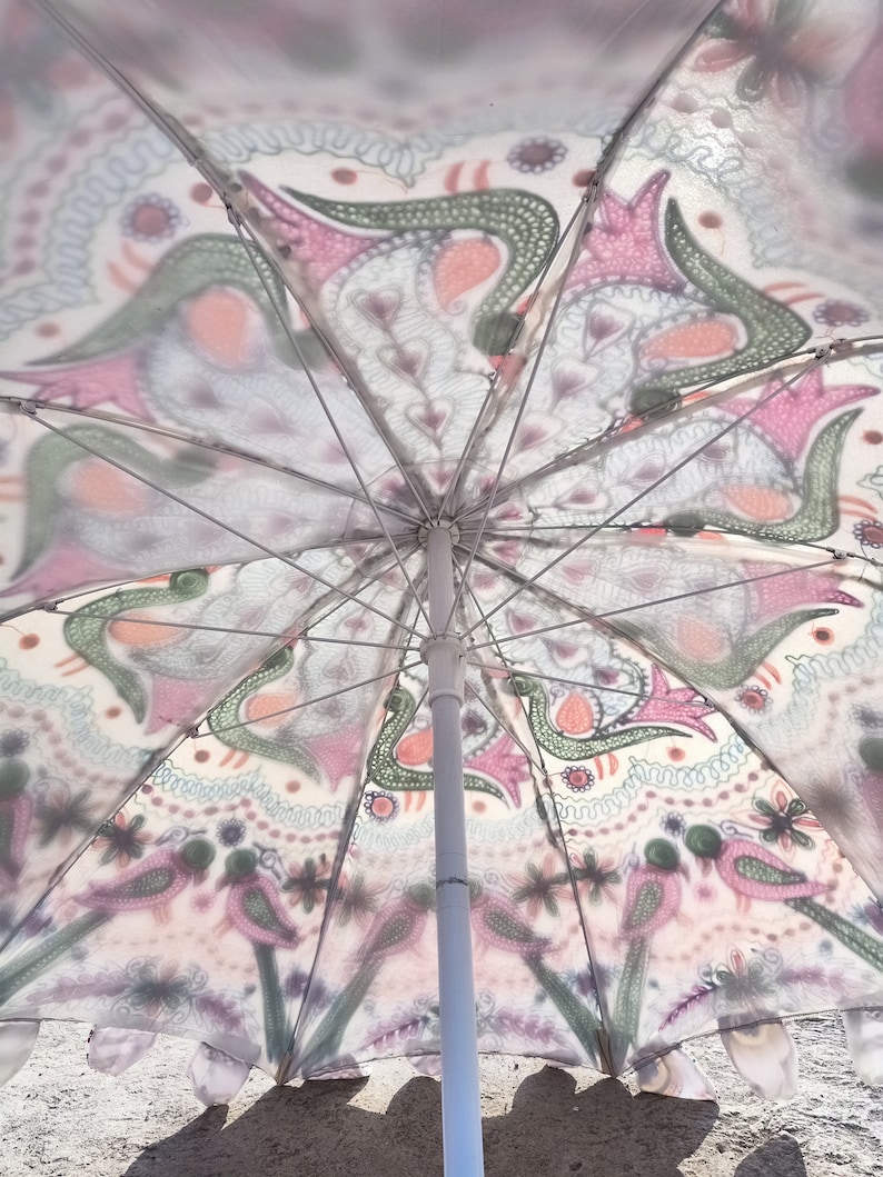 Może przedstawiać: Biały parasol plażowy z kolorowym, skomplikowanym wzorem. Wz&oacute;r zawiera kształty paisley, ptaki i elementy kwiatowe w odcieniach r&oacute;żu, zieleni i pomarańczy. Parasol ma biały słup i metalowe żebra.