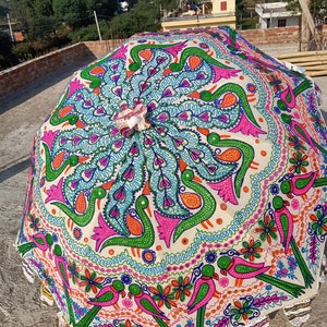 Może przedstawiać: Duży, otwarty, dekoracyjny parasol z żywym, ręcznie malowanym wzorem. Parasol ma kremowe tło z kolorowymi motywami pawia i ptak&oacute;w w odcieniach r&oacute;żu, zieleni, pomarańczy i niebieskiego. Krawędzie mają falisty brzeg.