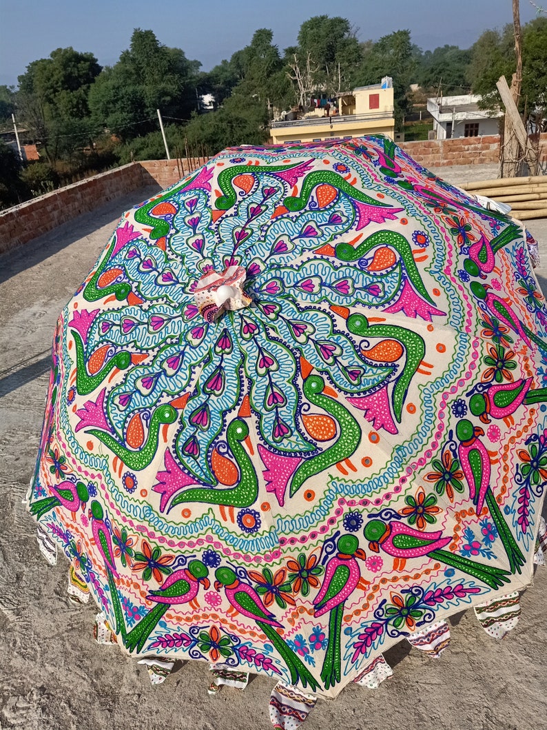 Może przedstawiać: Duży, otwarty, dekoracyjny parasol z żywym, ręcznie malowanym wzorem. Parasol ma kremowe tło z kolorowymi motywami pawia i ptak&oacute;w w odcieniach r&oacute;żu, zieleni, pomarańczy i niebieskiego. Krawędzie mają falisty brzeg.