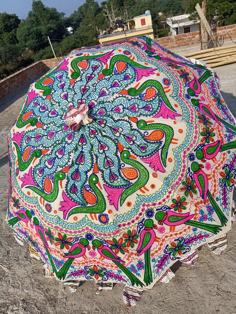 Może przedstawiać: Kolorowy, otwarty parasol z żywym, ręcznie malowanym wzorem. Parasol ma kremowe tło z powtarzającym się wzorem ptak&oacute;w, kwiat&oacute;w i wirujących linii w odcieniach r&oacute;żu, zieleni, niebieskiego i pomarańczowego. Krawędzie są ząbkowane.