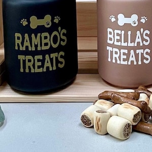 May include: Three ceramic dog treat jars with wooden lids. The jars are black, brown, and white. Each jar has a bone and paw print design and is labelled with a dog's name and "Treats". A pile of dog treats is in front of the jars.