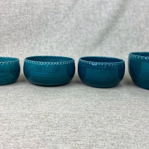 Può includere: Quattro ciotole in ceramica color turchese di diverse dimensioni con un bordo decorativo bianco. Le ciotole sono disposte in fila dalla più piccola alla più grande su una superficie testurizzata di colore grigio chiaro.