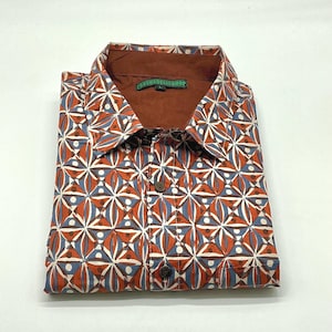 Può includere: Camicia button-down piegata con un motivo geometrico nei toni ruggine, blu e bianco. La camicia ha un colletto marrone e il testo "PSYCHEDELICSHOP" è visibile sul colletto.