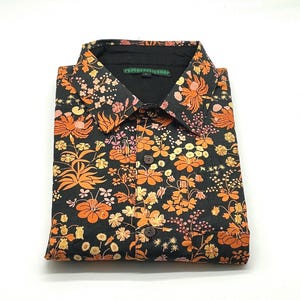 Könnte beinhalten: Schwarzes Hemd mit einem auffälligen Blumenmuster in Orange, Gelb und Rosa. Der Kragen ist schwarz und trägt den Schriftzug "PSYCHEDELICSHOP". Das Hemd ist sauber gefaltet.