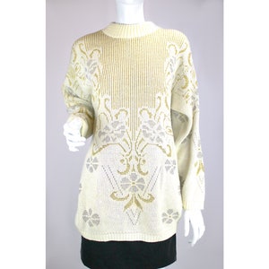 Pode incluir: Um suéter cor creme com um padrão floral dourado e prateado. O suéter tem um decote redondo e mangas compridas.