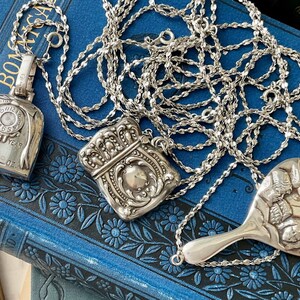 May include: Three silver necklaces with different pendants. The first pendant is a silver pocket watch with a face that reads "Month Year LBS OZ". The second pendant is a silver locket with an ornate design. The third pendant is a silver hand mirror with a floral design.