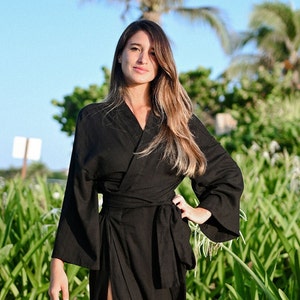 May include: A long, black wrap robe with wide sleeves and a waist tie. The robe has a high slit on the left side. The person is standing on a sandy beach with green plants in the background.