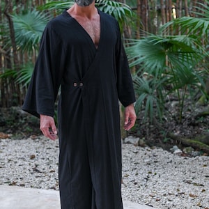 May include: A long-sleeved black robe featuring a V-neckline and a wrap-style front closure secured with two small brown buttons on the side. The garment has a loose, flowing silhouette that extends to the ankles with wide, straight sleeves.
