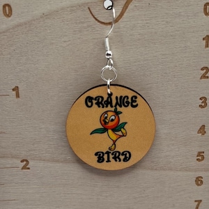 May include: A round wooden earring featuring a cartoon orange bird with green leaves and the text "ORANGE BIRD" in black. The earring hangs from a silver-toned metal hook on a light wood display card with printed inch and centimeter rulers.
