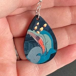 May include: A teardrop-shaped wooden earring with a black background and a blue and green cartoon image of a creature with large eyes and an open mouth. The earring has a silver jump ring and chain.