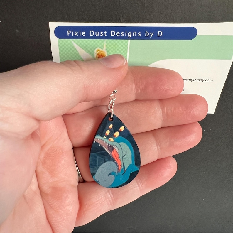 May include: A teardrop-shaped wooden earring with a black background and a blue and green cartoon image of a creature with large eyes and an open mouth. The earring has a silver jump ring and chain.