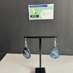 May include: A pair of teardrop-shaped earrings with a black background and a colourful image of a dolphin jumping out of the water. The earrings are silver and have a hook fastening.