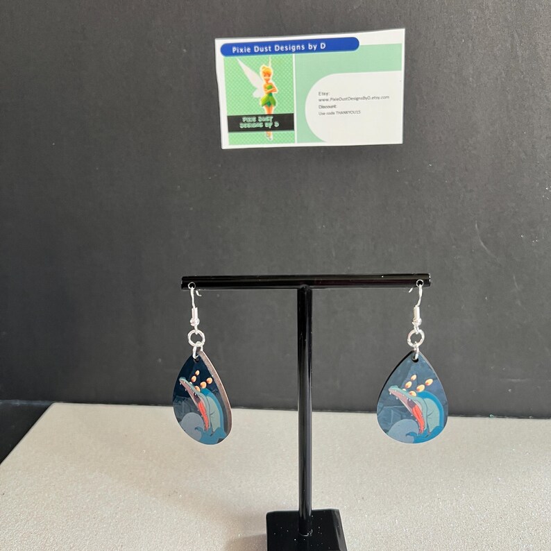 May include: A pair of teardrop-shaped earrings with a black background and a colourful image of a dolphin jumping out of the water. The earrings are silver and have a hook fastening.