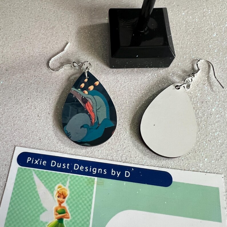 May include: A pair of teardrop-shaped earrings with a blue and grey cartoon image of a creature with large eyes and a wide open mouth. The other earring is blank and white.