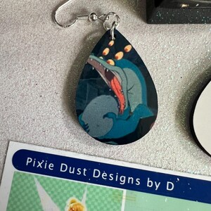 May include: A pair of dangle earrings featuring a blue and red cartoon monster with multiple eyes. The earrings are made of wood and have a teardrop shape. The earrings are hanging from silver hooks.