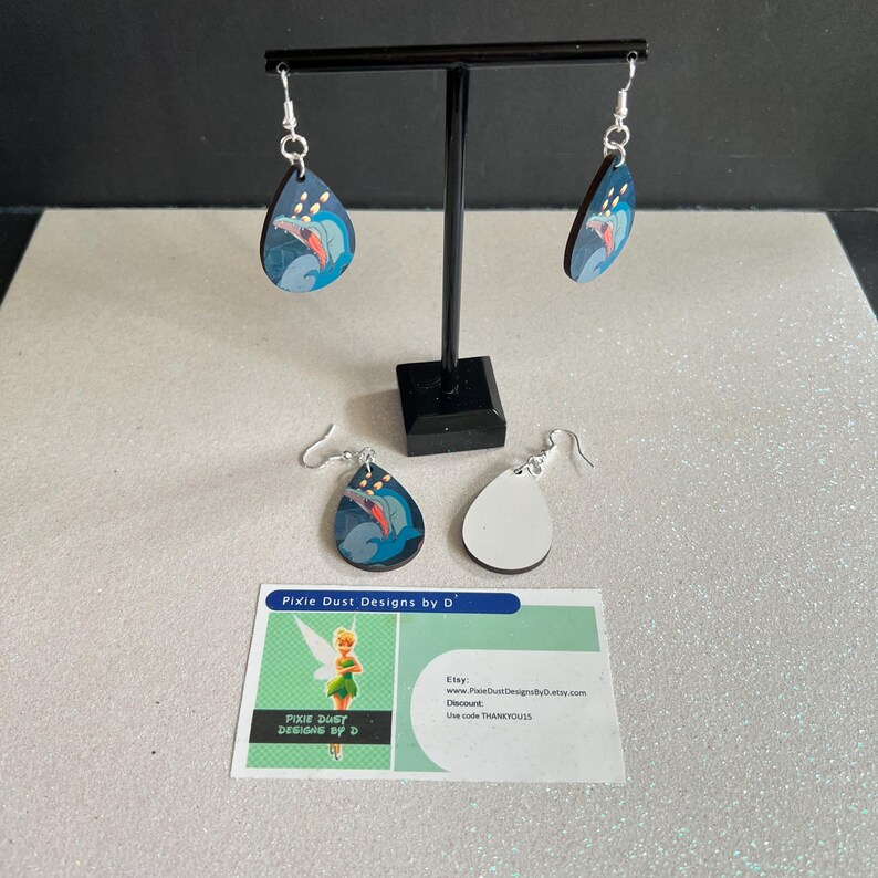 May include: A pair of teardrop-shaped wooden earrings featuring a blue and white image of a dolphin. The earrings are displayed on a black stand. A business card with the text "Pixie Dust Designs by D" is in the foreground.