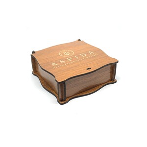 May include: A wooden box with a dark brown stain and a gold inscription that reads "ASPIDA  HANDMADE WITH PASSION & DETAIL".