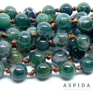 May include: A close-up of a green agate gemstone beaded necklace with brown cord. The beads are round and have a mottled green and white pattern.