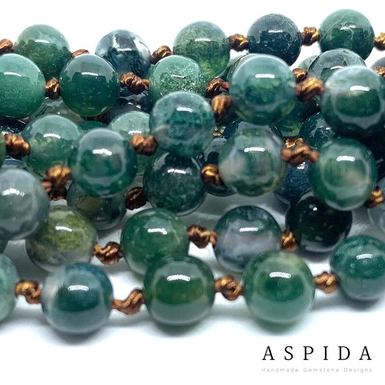 May include: A close-up of a green agate gemstone beaded necklace with brown cord. The beads are round and have a mottled green and white pattern.