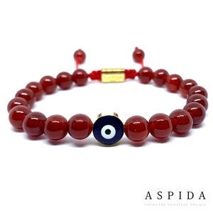 Peut inclure: Bracelet en perles de cornaline rouge avec un charme œil maléfique en or. Le charme a un œil bleu avec une pupille blanche et une bordure dorée.