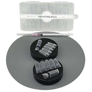 May include: A clear plastic box labeled "Square Nail Tips" and "AQUARIUM NAILS" contains numerous clear nail tips. Below, clear nail tips are arranged on black circular platforms. A syringe is also present.