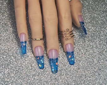 May include: Long, blue glitter nail tips with a clear base and a thin line of blue glitter at the base of the nail. The nails are decorated with small, silver stars and moons.