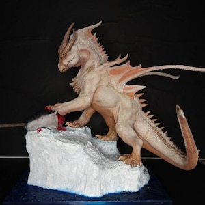 May include: A white dragon figurine with red accents, standing on a white ice base with a blue water base. The dragon is holding a narwhal in its claws.