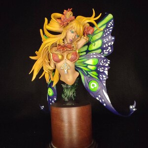 May include: A sculpted bust of a fairy with long blonde hair, wearing a red and gold bodice and a green and purple butterfly wing. The fairy is standing on a wooden base.
