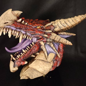 May include: A red dragon head sculpture with a purple tongue and detailed scales. The dragon's horns are curled and the sculpture is painted with a realistic finish.