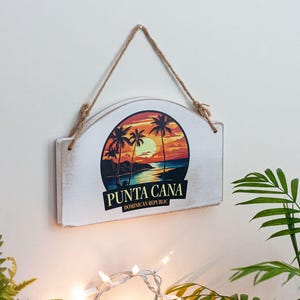 A white wooden sign with a tropical beach scene and palm trees is hanging on a wall, surrounded by greenery and a string.