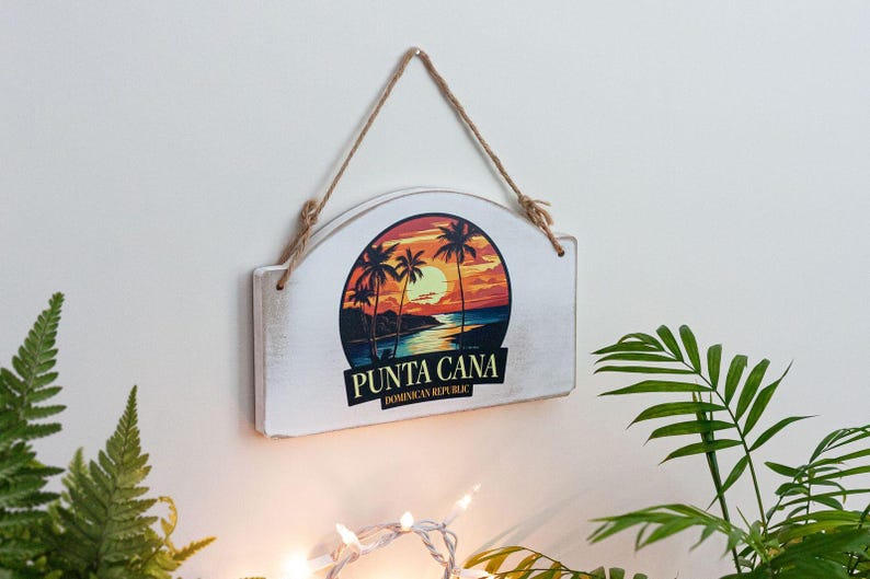 A white wooden sign with a tropical beach scene and palm trees is hanging on a wall, surrounded by greenery and a string.