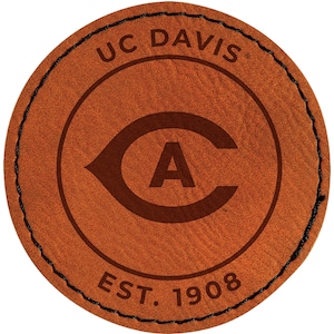 May include: A round, brown leather patch with the text "UC DAVIS" arched across the top. Inside a circle is a stylized "C" with an "A" in the center. The text "EST. 1908" is at the bottom. The edge is stitched with black thread.