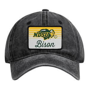 May include: A charcoal grey baseball cap with a rectangular patch on the front. The patch features a yellow and white striped background with a green NDSU logo, a graphic of a bison head, and the word Bison written in a green cursive script.