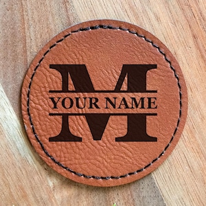 May include: A round, tan leather coaster with a stitched border. The coaster features a large, dark brown letter "M" above the words "YOUR NAME" and another "M" below. The design is centered on the coaster.