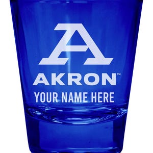 May include: A cobalt blue glass shot glass featuring a white printed design. The design includes a large letter A, the word AKRON, and the text YOUR NAME HERE. The glass has a tapered cylindrical shape with a thick, clear base and smooth walls.