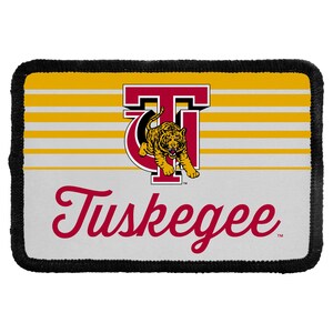May include: A rectangular patch with a black stitched border. The design features horizontal gold and white stripes, a maroon letter T logo with a gold tiger graphic, and the word Tuskegee written in a maroon cursive script font below the logo.