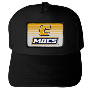 May include: A black trucker-style baseball cap featuring a rectangular patch on the front. The patch displays a yellow letter C above the word MOCS in bold white letters with a dark blue outline, set against a background of horizontal yellow lines.