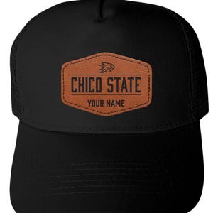 May include: A black trucker hat featuring a brown faux leather patch on the front. The patch is hexagonal and displays a wildcat logo above the text CHICO STATE and the words YOUR NAME in black lettering, with a mesh back and solid front panel.