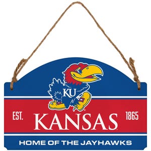 May include: A wooden sign with a blue and red design featuring the text "KANSAS" and a Jayhawk mascot. The sign includes the text "EST. 1865" and "HOME OF THE JAYHAWKS". A brown rope is attached for hanging.