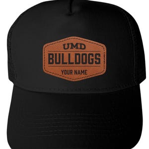 May include: A black trucker hat featuring a brown faux leather patch on the front. The patch is hexagonal and displays the black text UMD, BULLDOGS, and YOUR NAME. The hat has a solid front panel and a mesh back panel with a curved brim.