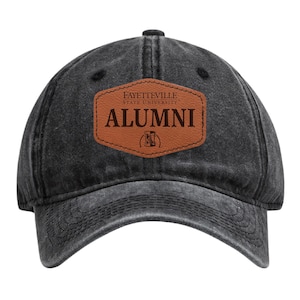 May include: A black, distressed baseball cap with a curved brim. The cap features a brown leather patch with the words "FAYETTEVILLE STATE UNIVERSITY ALUMNI" in a serif font. The patch also includes a university logo.