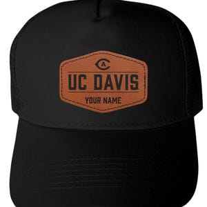 May include: A black trucker hat featuring a brown, hexagonal, leather-like patch on the front. The patch displays the UC Davis logo, the text UC DAVIS, and the words YOUR NAME in black lettering. The hat has a solid front panel and mesh back.