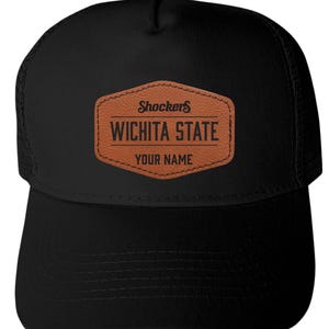 May include: A black trucker hat featuring a brown faux leather patch on the front. The patch is hexagonal and displays the text Shockers, WICHITA STATE, and YOUR NAME in black lettering. The hat has a solid front panel and mesh back panels.