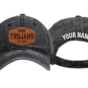 May include: A black washed-denim baseball cap featuring a brown leather-look patch on the front with the text TROY TROJANS EST. 1887. The back of the cap displays the white text YOUR NAME and includes an adjustable metal buckle strap closure.